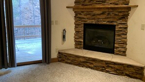 Une cheminée en pierre avec une cheminée décorative, une terrasse en bois visible à travers une porte-fenêtre coulissante et un plancher recouvert de moquette.