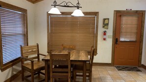 Un espace repas avec une table et des chaises en bois, deux lampes suspendues, des stores en bois, un extincteur et une porte portant l’inscription « SORTIE ».
