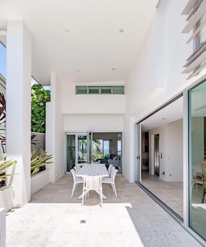 A modern outdoor patio with a dining table and chairs, a glass door leading to a living area, and a view of greenery.