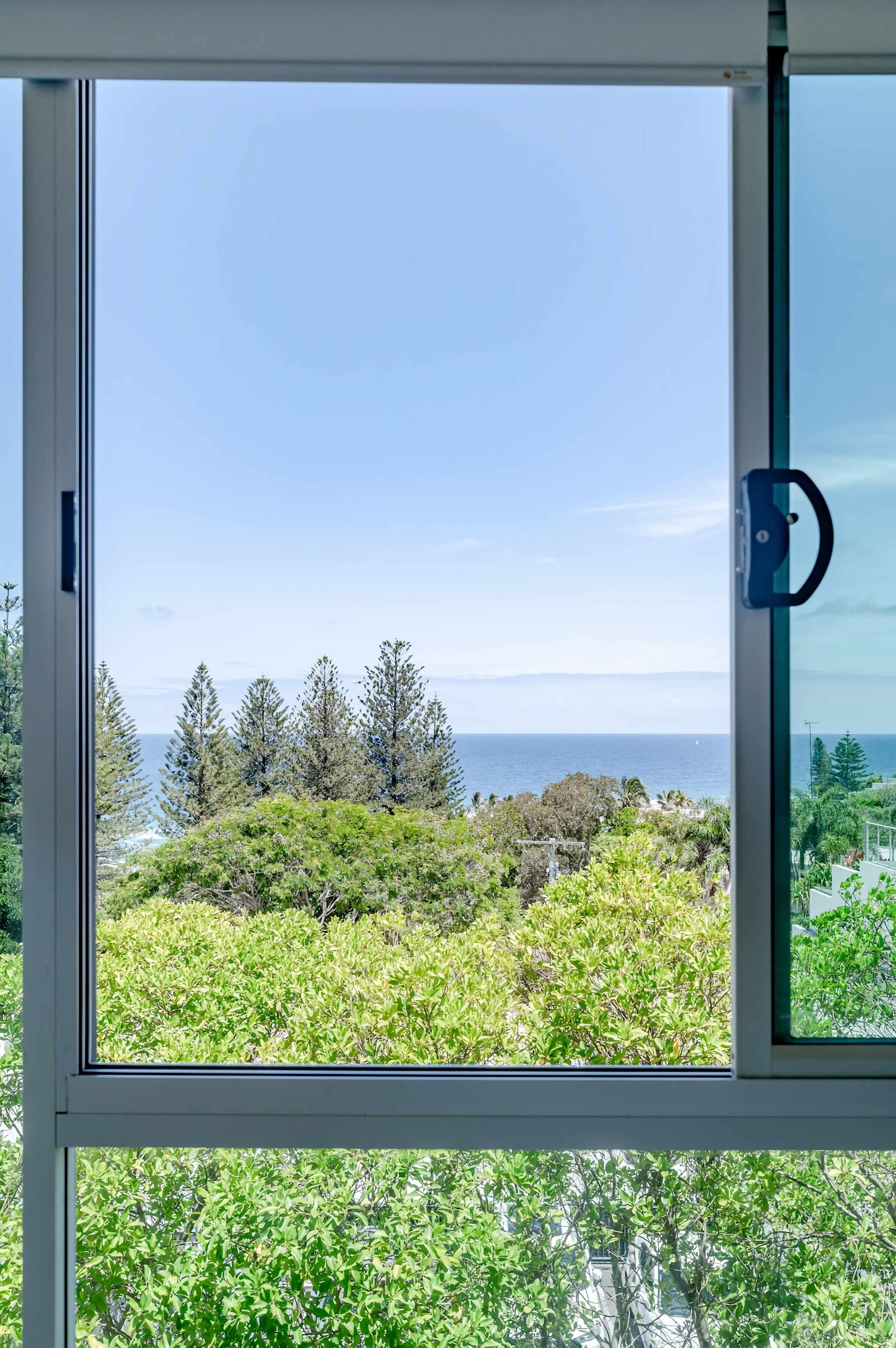 Une fenêtre donnant sur un paysage côtier, avec des arbres et l’océan en toile de fond.