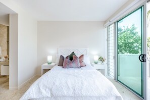 A modern bedroom with a large bed, two bedside tables with lamps, a white headboard, and a sliding glass door leading to a balcony.