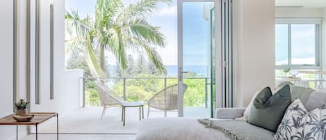 A modern bedroom with a large sliding glass door leading to a balcony with a view of palm trees and the ocean.