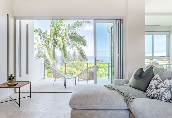 A modern bedroom with a large sliding glass door leading to a balcony with a view of palm trees and the ocean.