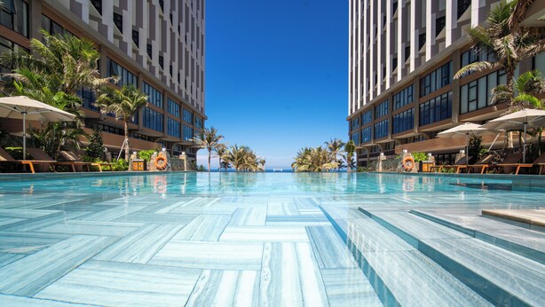 Una piscina con suelo de baldosas, rodeada de sillas de descanso y sombrillas, con edificios altos al fondo.