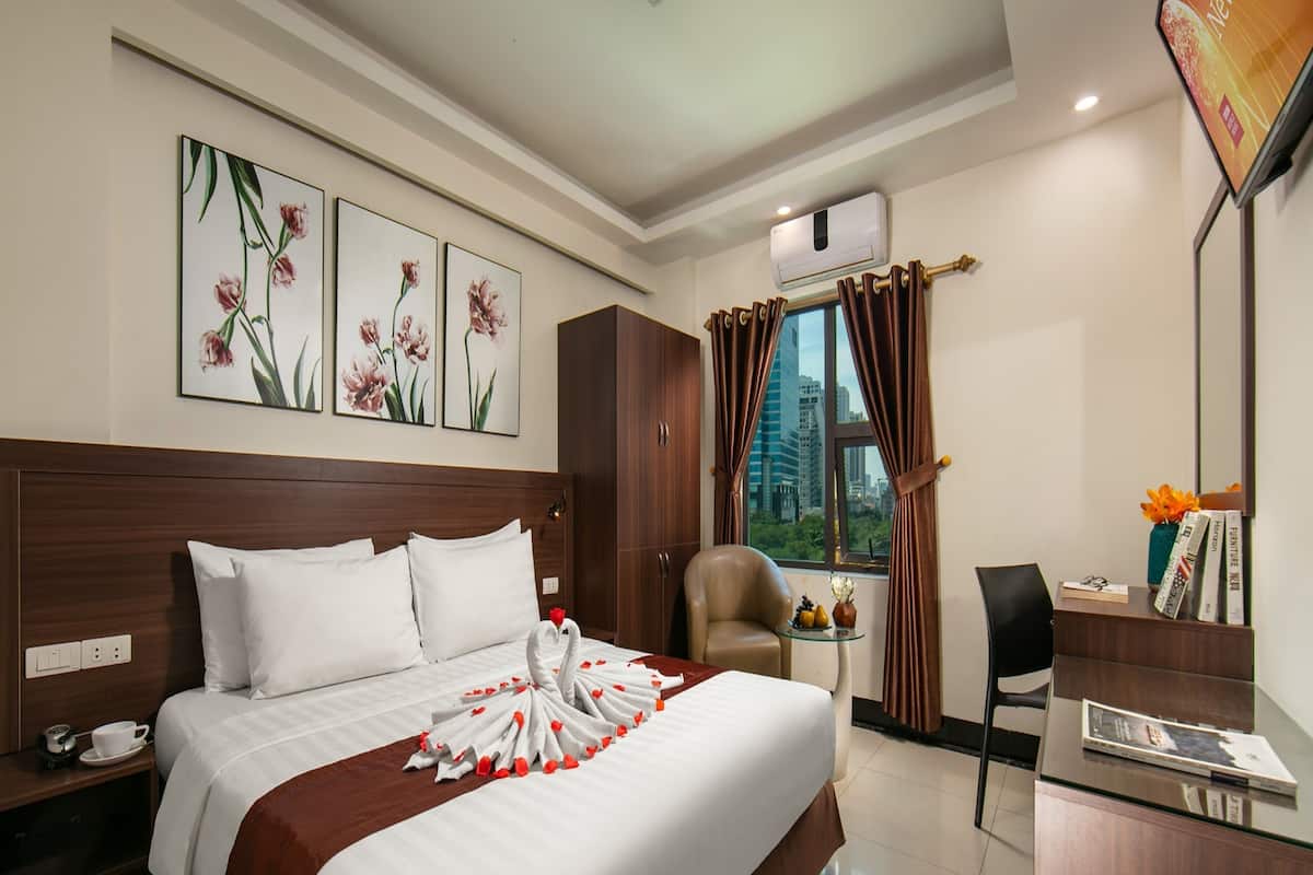 A hotel room with a large bed, a desk, a chair, a window with curtains, and artwork on the wall.