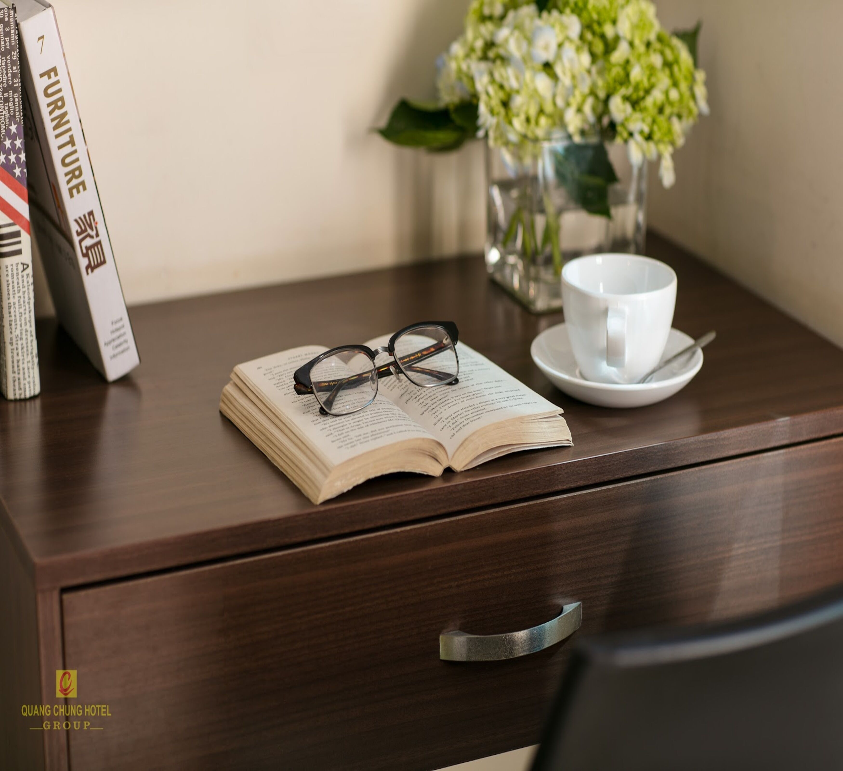 A wooden desk with an open book, a pair of glasses, a white cup, and a vase with flowers.