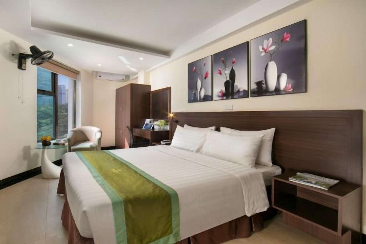 Quarto de hotel com uma cama grande, uma escrivaninha com cadeira, mesa de cabeceira e obra de arte na parede.