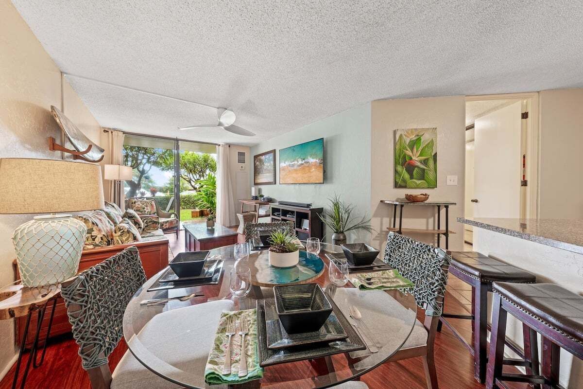 A spacious dining area with a round table, chairs, and a ceiling fan. There are plants and artwork on the walls, and a view of the outdoors through large windows.