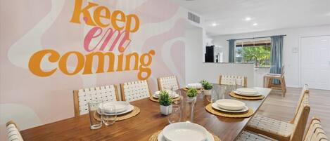 A dining area with a long wooden table set for six, white chairs, and a kitchen visible in the background.