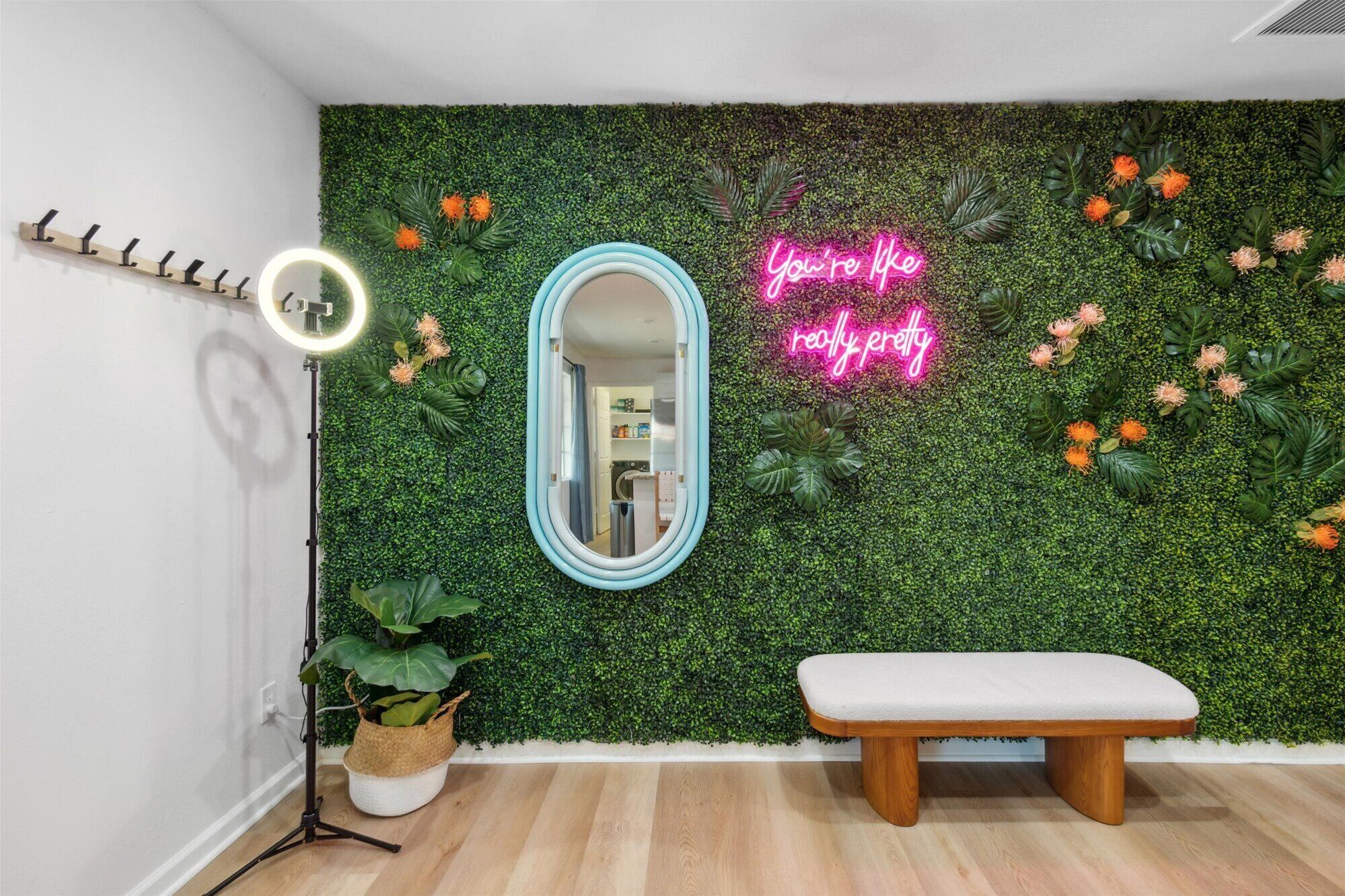 A room with a green wall covered in foliage and flowers, a neon sign, a mirror, a bench, and a ring light.