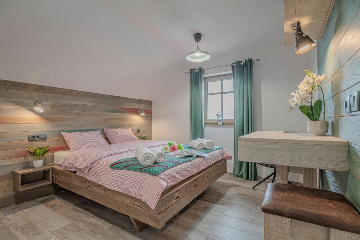 Une chambre à coucher avec un lit en bois, des tables de chevet, un petit bureau, une fenêtre avec des rideaux et un luminaire suspendu.