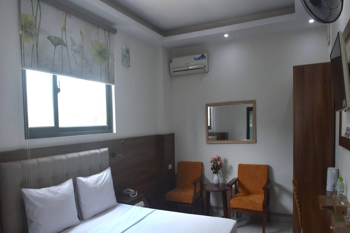 A hotel room with a bed, two chairs, a small table with a vase, an air conditioning unit, and a mirror on the wall.