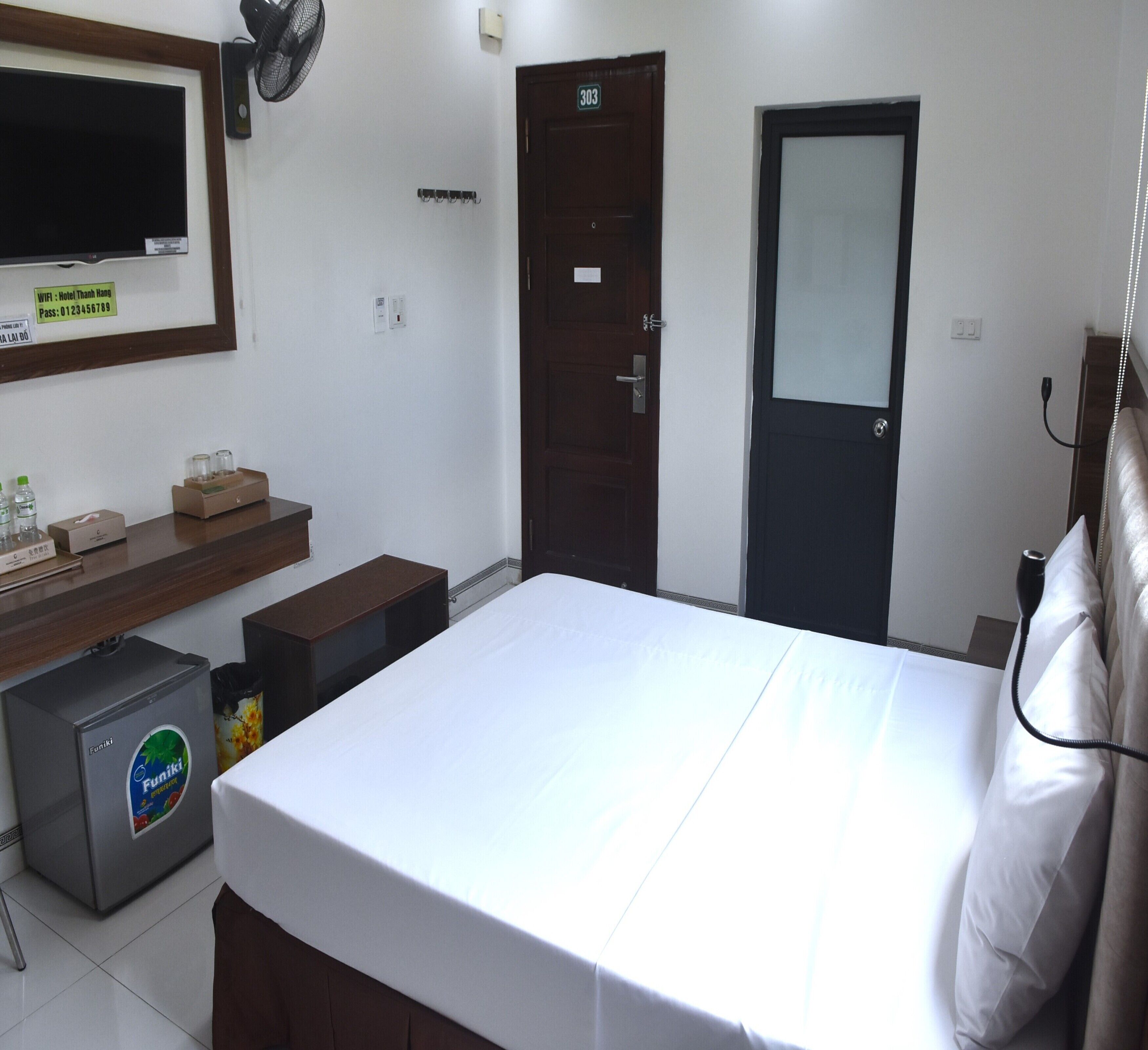 A hotel room with a bed, a desk with a computer, a minibar, a TV, and a door with a number.