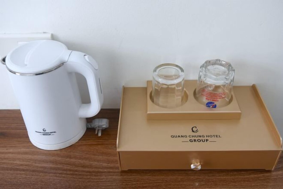 A white electric kettle and a glass carafe on a wooden table, with a brown box labeled "QUANG CHUNG HOTEL GROUP" in the background.