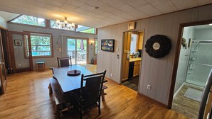 A dining area with a wooden table and chairs, a kitchen visible through an open door, and a large window with a view of trees.