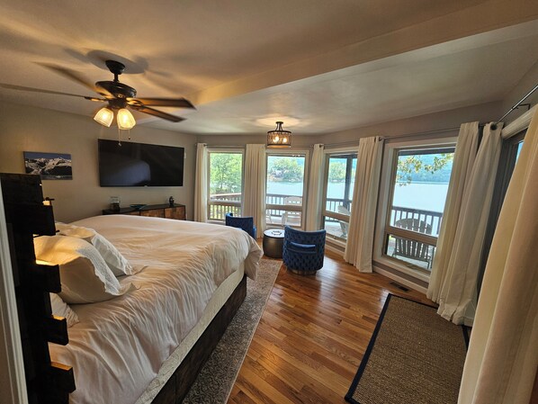 A bedroom with a large bed, a ceiling fan, a TV, and a view of a lake through large windows.