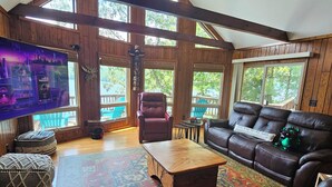 A spacious living room with a high ceiling, wooden beams, large windows, a leather sofa, a coffee table, and a television.