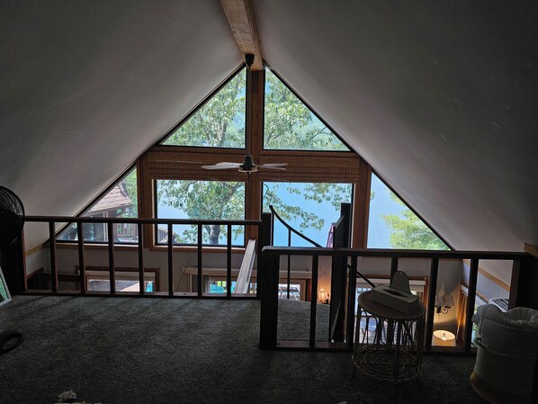 Habitación en ático con una ventana grande, ventilador de techo y vistas a una casa y árboles.