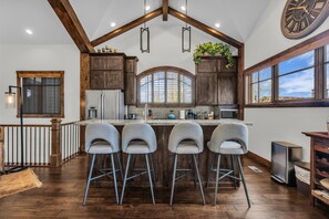 A kitchen with wooden cabinets, a large island with bar stools, a clock, and a window with a view.