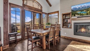 A dining area with a wooden table and chairs, large windows with a mountain view, a fireplace, and a bookshelf. - Luxurious 4BD Near Canyons Village (Park City)