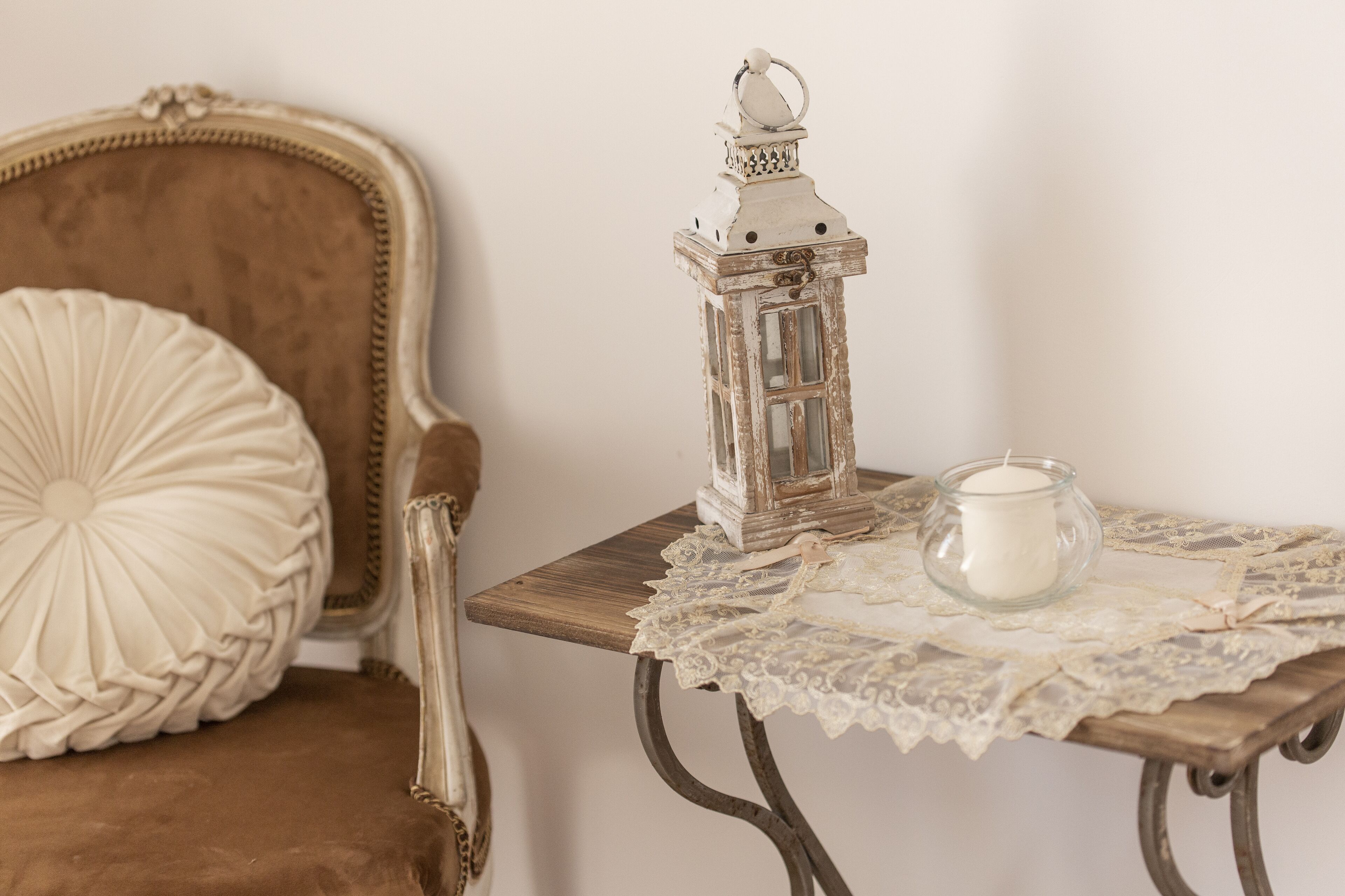 Uma cadeira antiga com uma almofada decorativa, uma mesa pequena com toalha de renda, uma vela e um candeeiro.