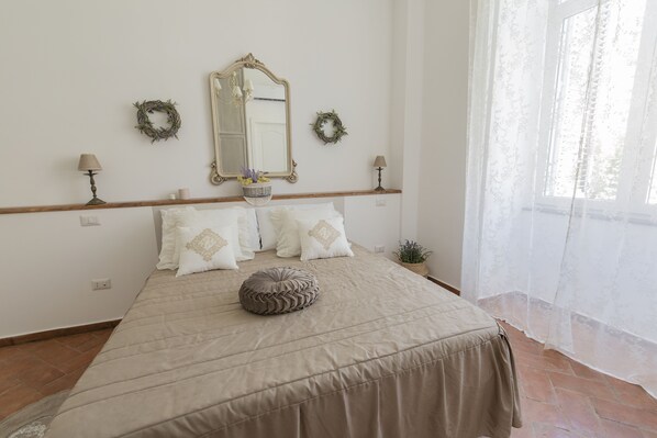 Un dormitorio ordenado con una cama grande, un espejo, dos apliques de pared y una ventana con cortinas translúcidas.