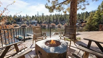 Terraza exterior con un hogar a fuego de piedra, sillas de madera y vista a un lago y zona boscosa.