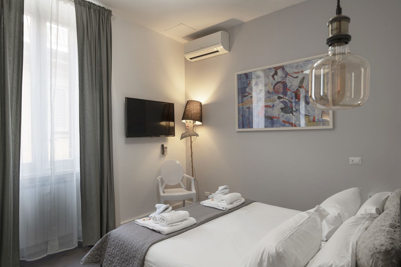 A hotel room with a bed, a chair, a TV, an air conditioner, and a painting on the wall.
