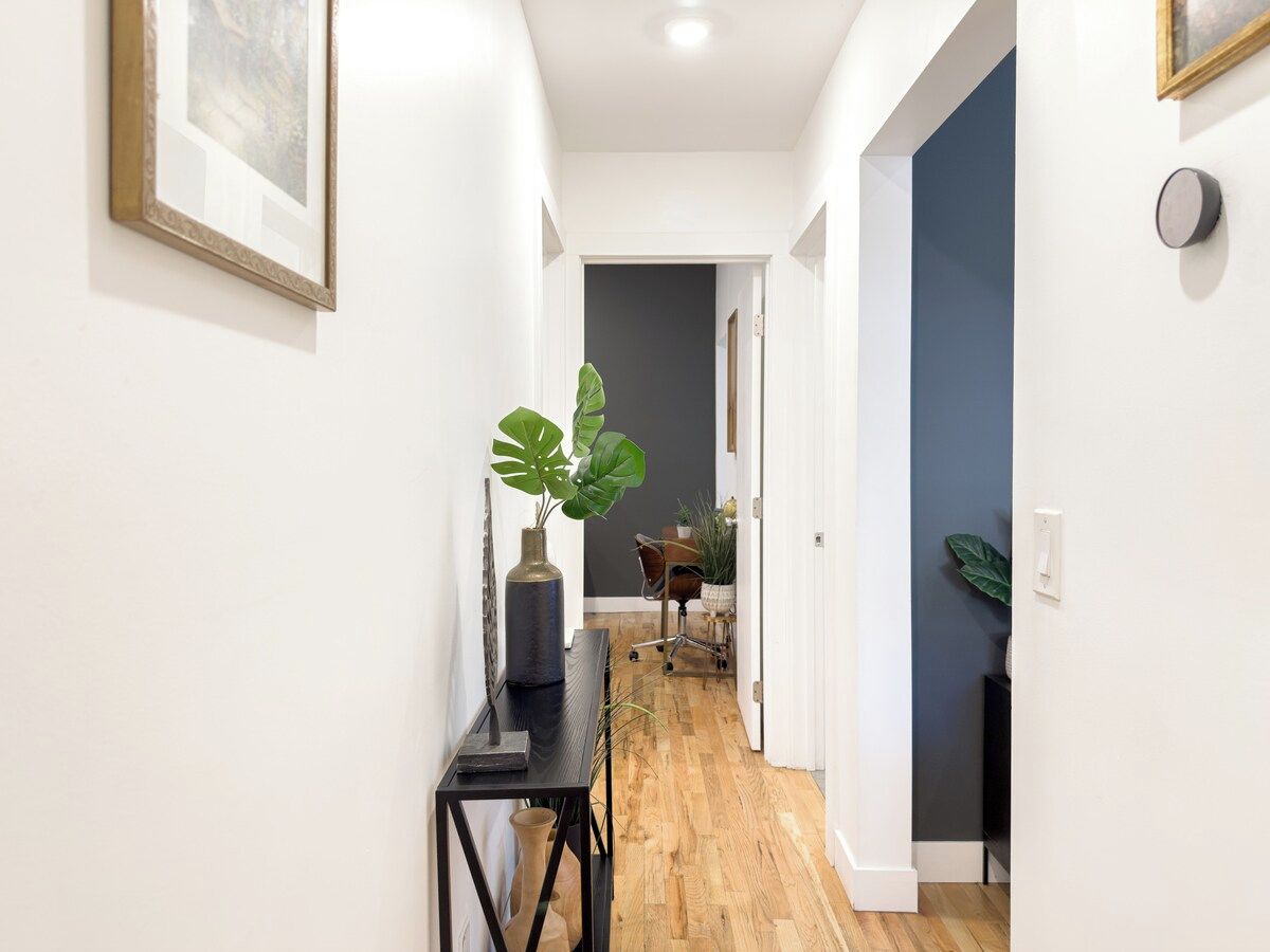 A hallway with a wooden floor, a black console table with a plant, and a door leading to a room with a dark wall.