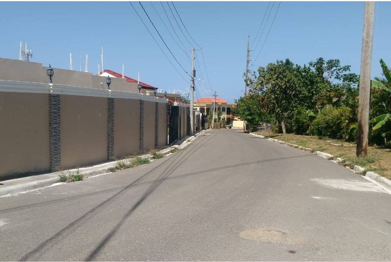 Una calle residencial tranquila con una casa moderna, techo rojo, pared de piedra y cielo despejado.