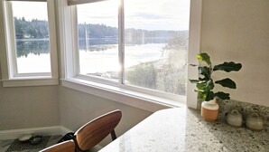 A kitchen with a large window overlooking a lake, a dining area with a table and chairs, and a potted plant on the counter. - 180' private beach and spacious entertainment oasis on Hammersley Inlet for 8! (Shelton)