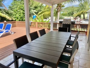 An outdoor patio with a long table and chairs, a grill, and a view of lush greenery. - Tropical Caribbean Oasis (Jolly Harbour)
