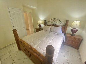 Un dormitorio con una cama de madera, dos mesitas de noche con lámparas, una puerta y piso de baldosas.