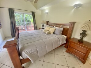 A bedroom with a large bed, a wooden headboard, a nightstand with a lamp, and a view of the outdoors through a window.