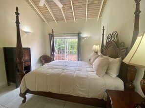 A bedroom with a large bed, a wooden headboard, a nightstand, a lamp, a chair, and a window with curtains. - Tropical Caribbean Oasis (Jolly Harbour)