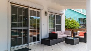 A covered patio area with a sofa and coffee table, large sliding glass doors, and a view of a blue house in the background.