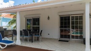 Un patio techado con una mesa y sillas, una piscina visible a través de un ventanal corredero y una parrilla pequeña al aire libre.
