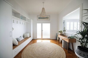 A room with a wooden floor, a woven rug, a bench with cushions, a wooden desk, a potted plant, and a door leading outside. - Sea Cliff Sands Gerroa (Gerroa)