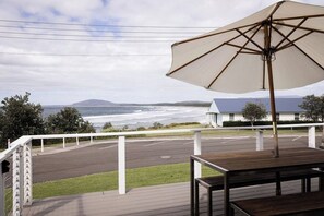 A deck with a table and an umbrella, overlooking a parking area and a beach with a building in the distance.