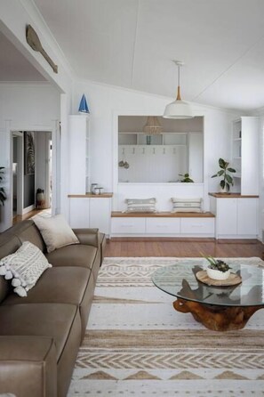 A modern living room with a glass coffee table, a sofa, and a kitchen in the background. - Sea Cliff Sands Gerroa (Gerroa)