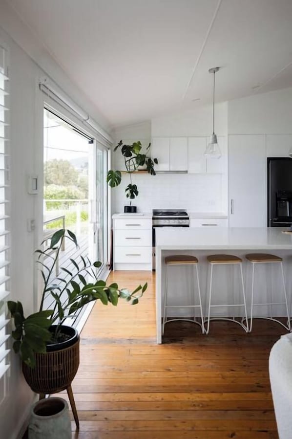 A modern kitchen with white cabinetry, a central island with stools, and a large window providing natural light. - Sea Cliff Sands Gerroa (Gerroa)