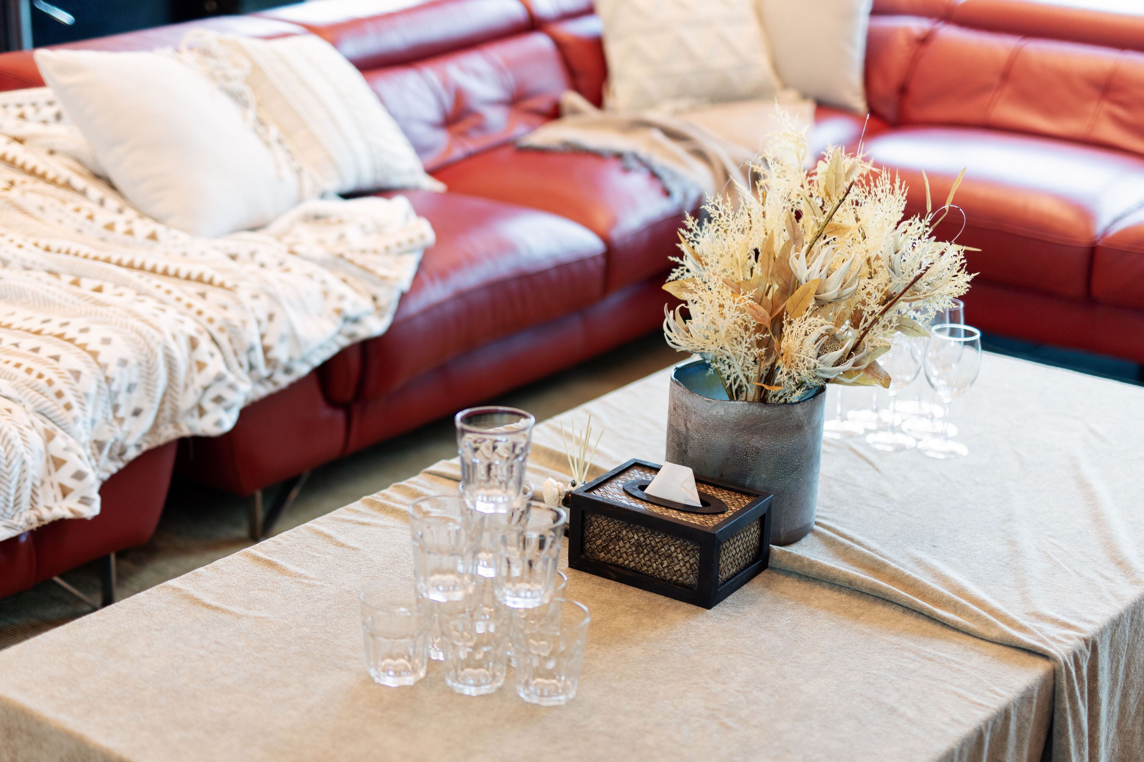 Una habitación con un sofá de cuero rojo, una mesa con un jarrón de flores secas y varios vasos.