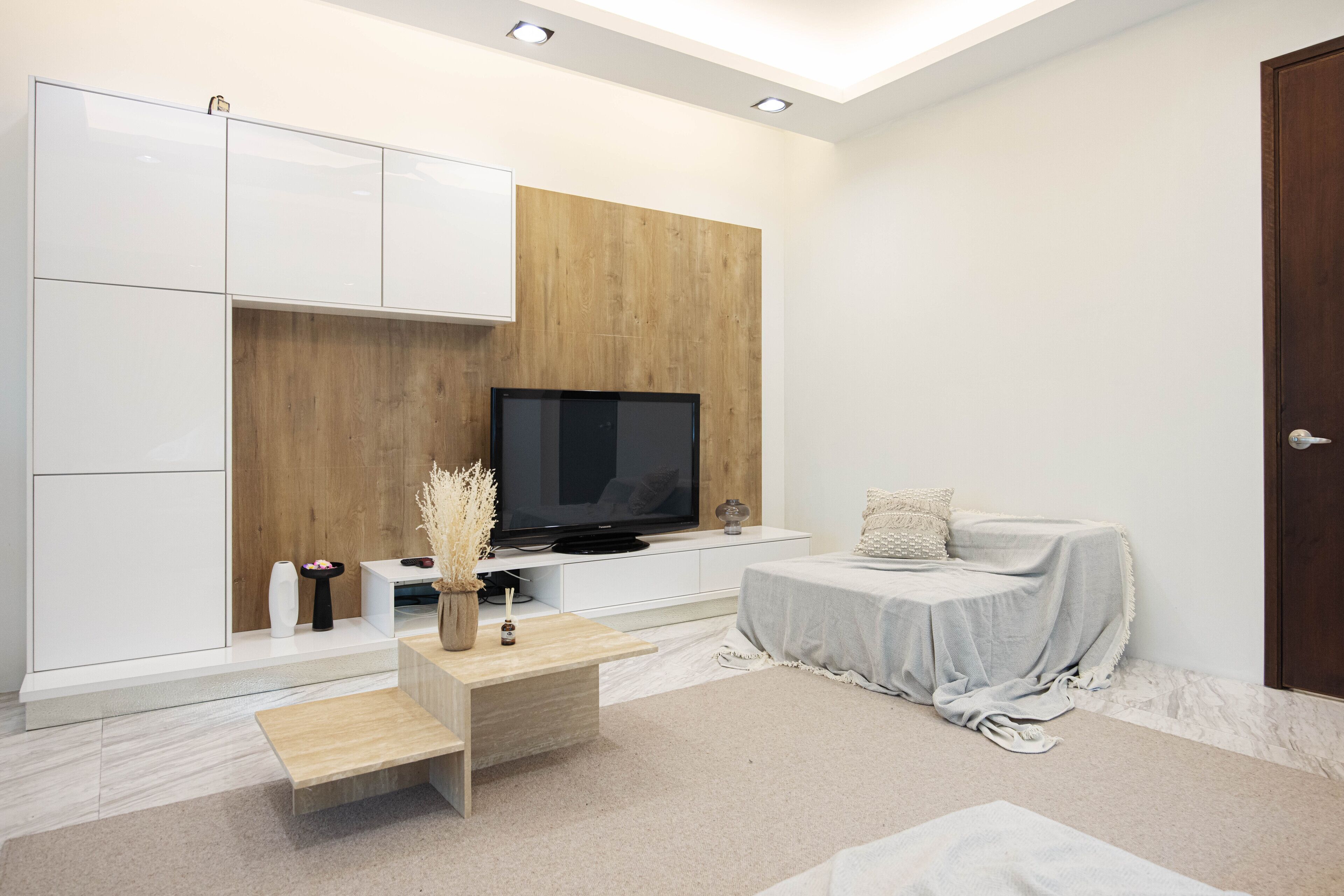 A modern living room with a flat-screen TV, white cabinetry, a wooden accent wall, a small coffee table, and a bed with a grey cover.