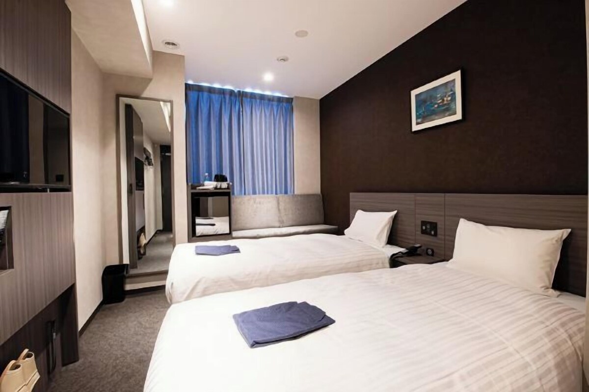 A hotel room with two beds, a TV, a desk, and a window with curtains.