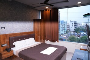 A hotel room with a bed, a desk, a telephone, and a window with a view of buildings and parked cars. - Vaidh Business Hotel (Hyderabad)