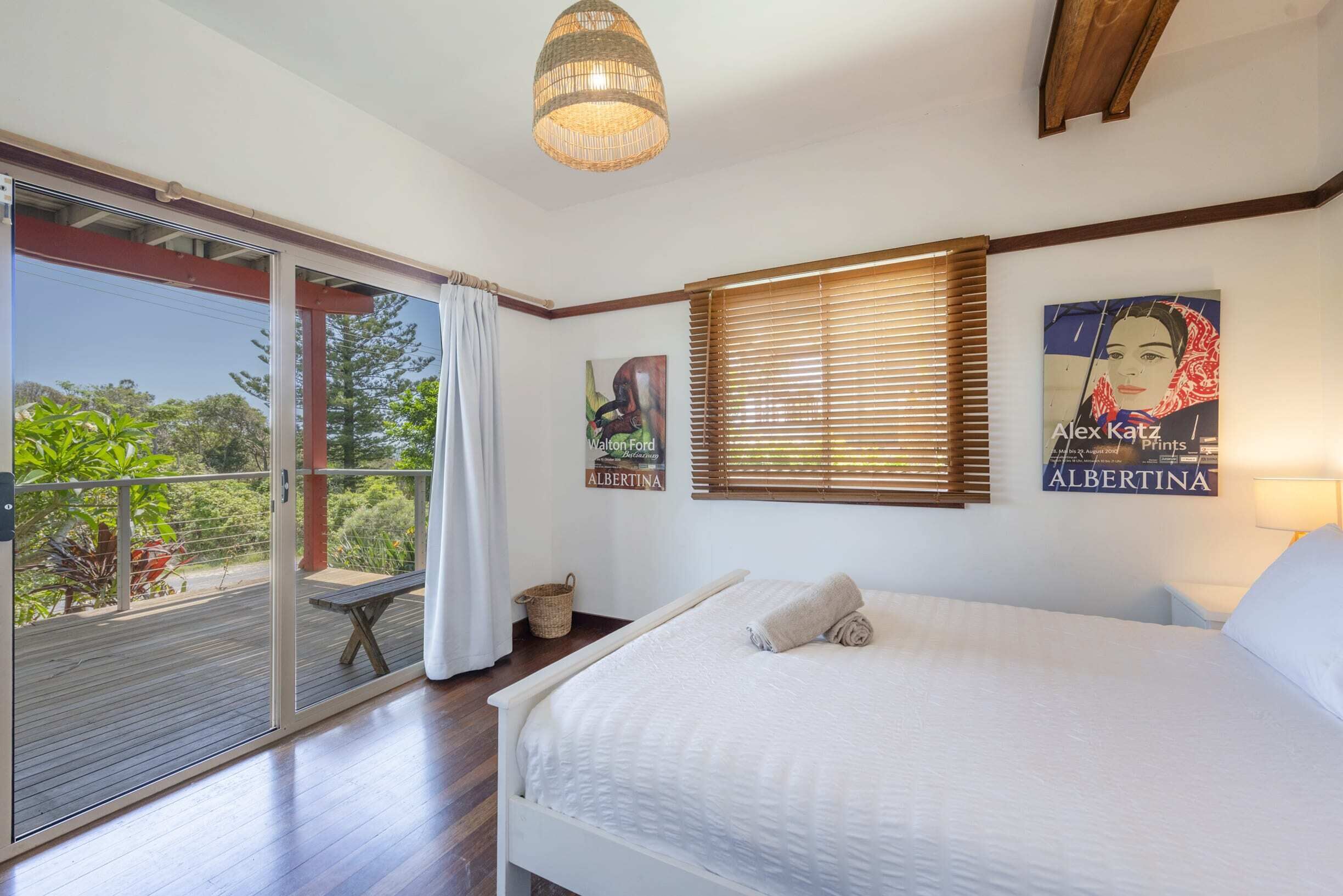 A bedroom with a bed, a window with blinds, a wall-mounted artwork, a ceiling light, and a sliding glass door leading to an outdoor area.