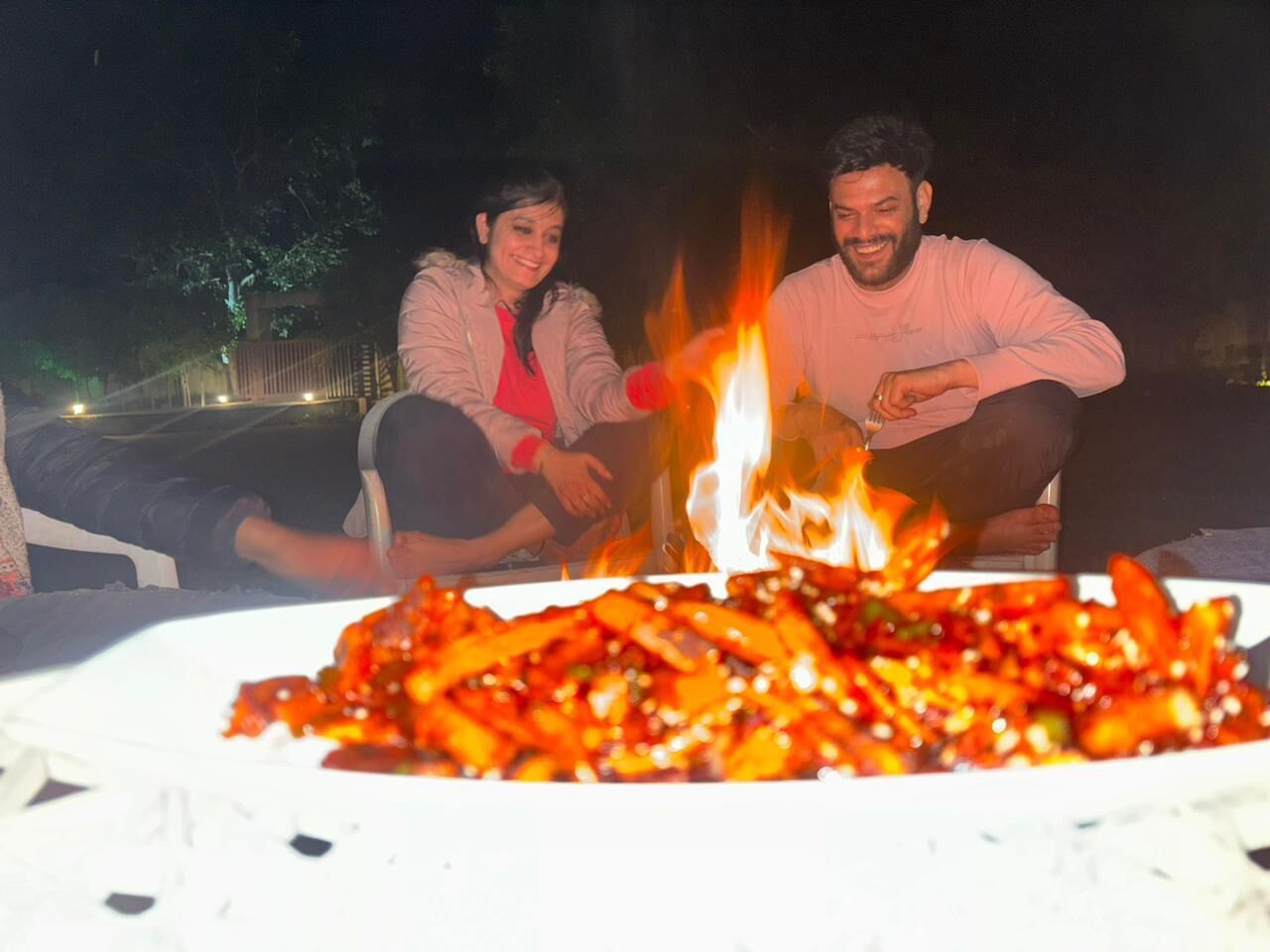 Two people sitting by a campfire with a plate of food in front of them.