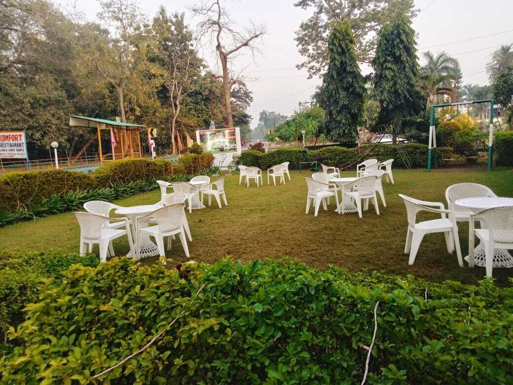 Un área de estar al aire libre con sillas y mesas de plástico blanco dispuestas sobre un césped, rodeada de follaje y árboles.