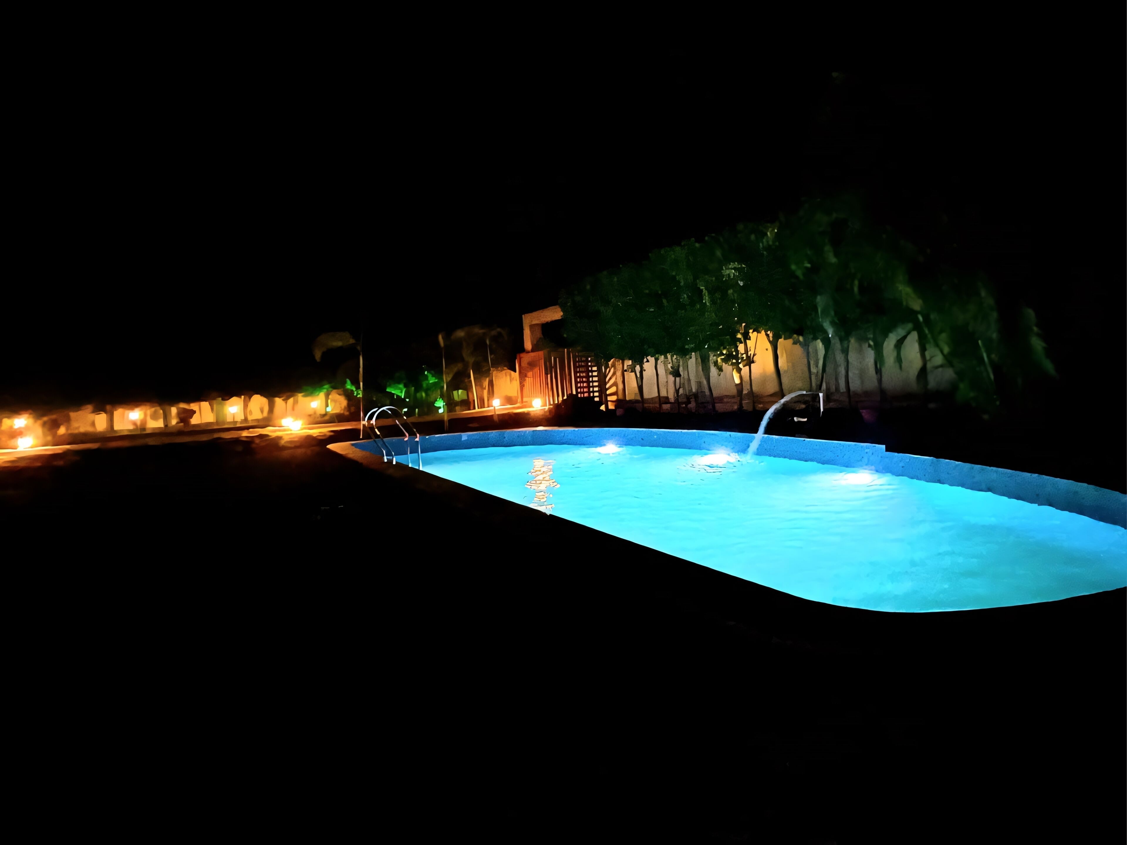 Piscina iluminada con luz ambiental, rodeada de árboles y con una fuente de agua.