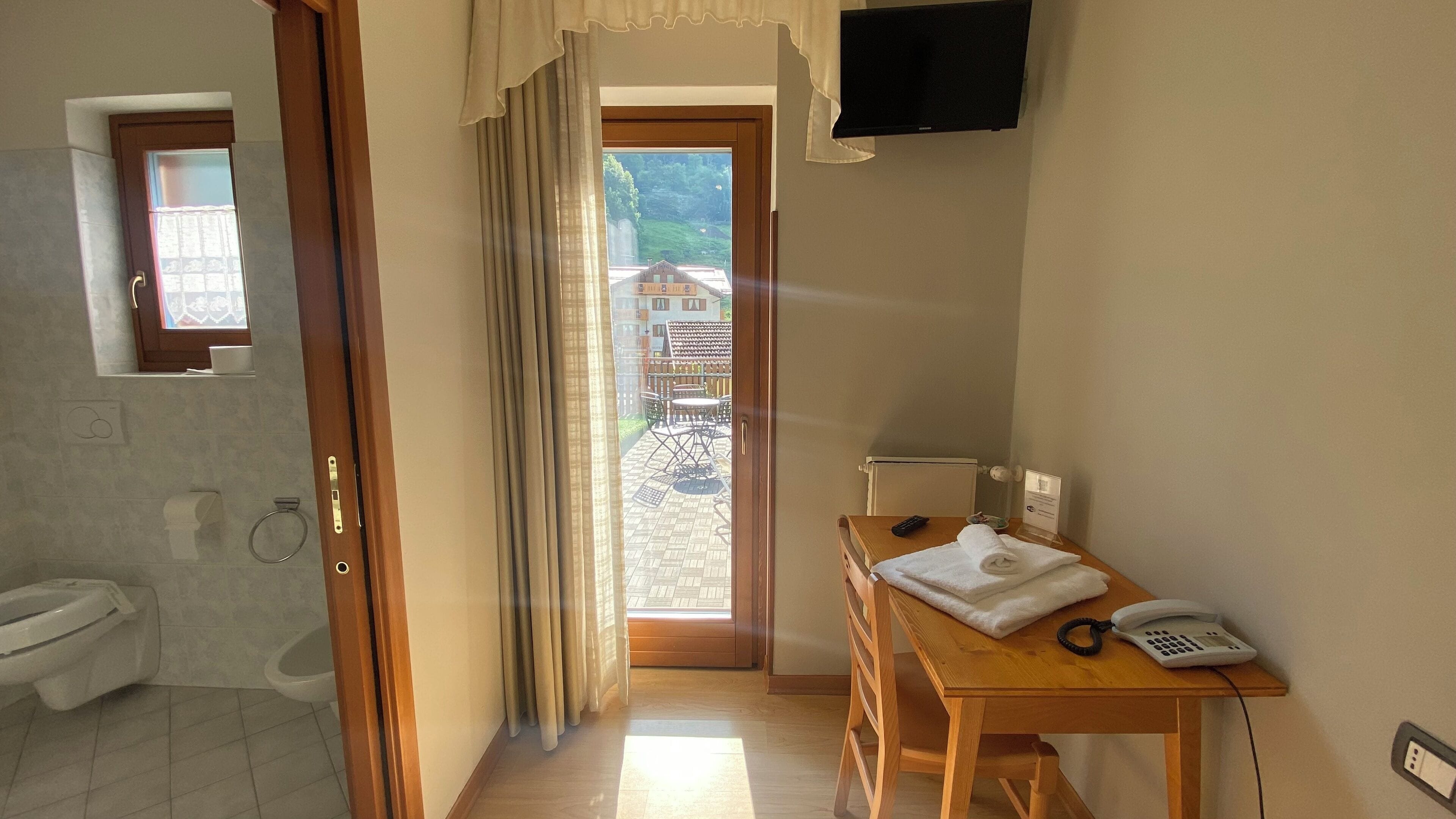 A hotel room with a bathroom, a wooden table with a telephone and a towel, and a view of a building through a window.